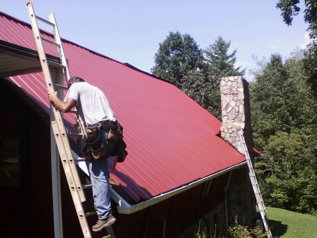 Roofer on a ladder beside a red metal roof and stone chimney — Soffit & Fascia Repair by Roofing Brothers in Fairland